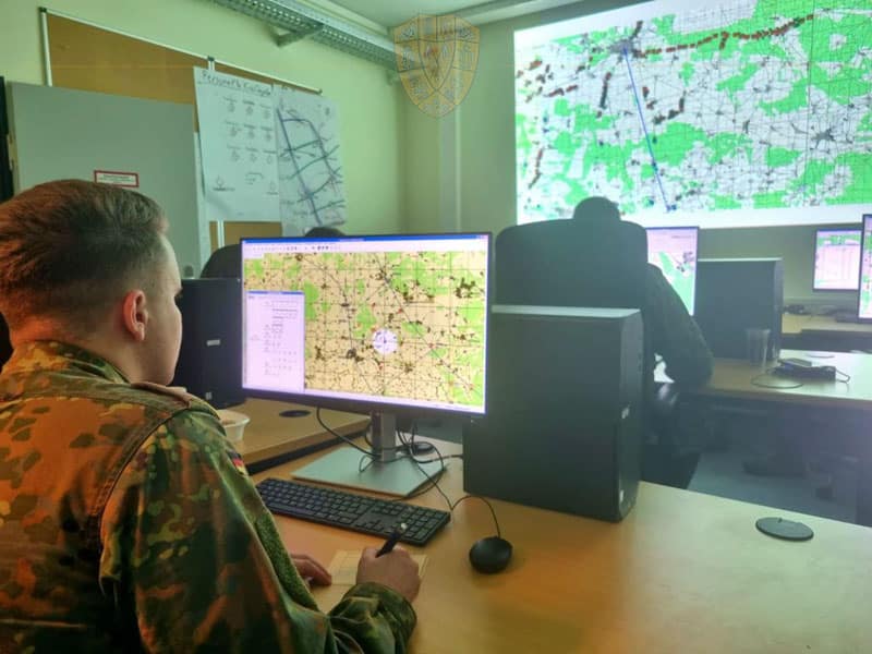 SOLDAT SITZT VOR DEM MONITOR MIT auf dem eine taktische Karte zu sehen ist. Im Hintergrund weitere Soldaten, ebenfalls for dem Computerbildschirm