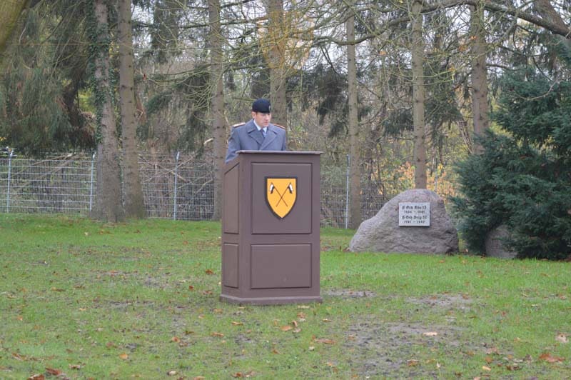 Der Kommandeur steht vor am Rednerpult und hält eine Ansprache. Das Rednerpult ziert das Bataillonswappen. Im Hintergrund das Ehrenmal.